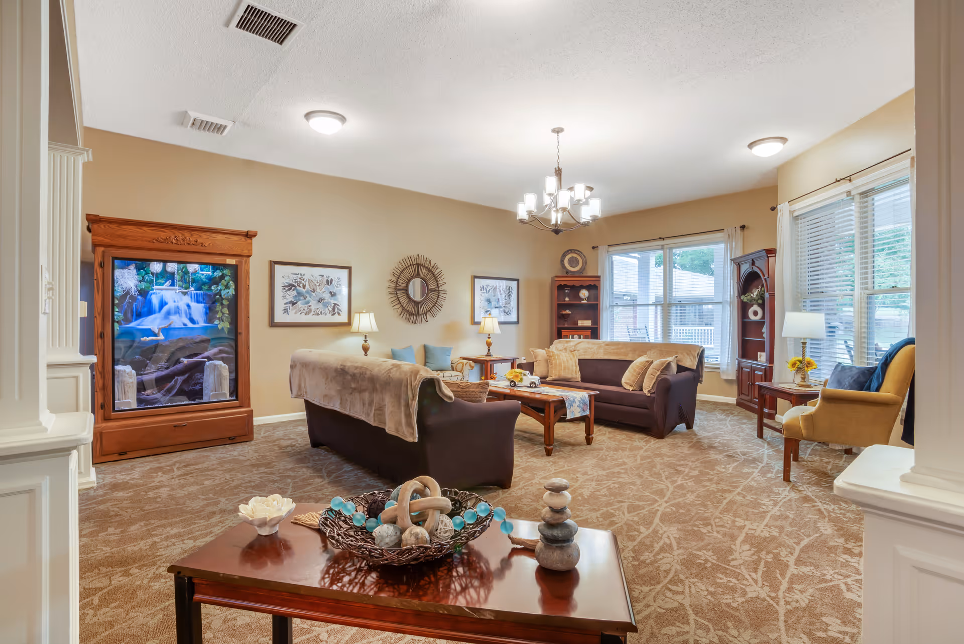 A cozy living room with two dark brown sofas, a wooden coffee table, and a yellow armchair near a window with white blinds. The room features beige walls, carpeted floor with a leaf pattern, a chandelier, and a large wooden cabinet with a waterfall aquarium. Decorative items and framed artwork adorn the walls and shelves.