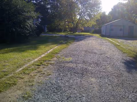 Gravel driveway leads to a senior living facility with trees and a small outbuilding.
