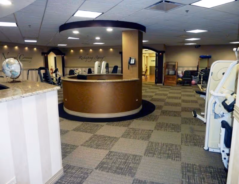 Interior view of a fitness or rehabilitation room at Norwood Crossing with exercise equipment, a circular reception desk in the center, a globe on a counter to the left, and beige walls with motivational text.
