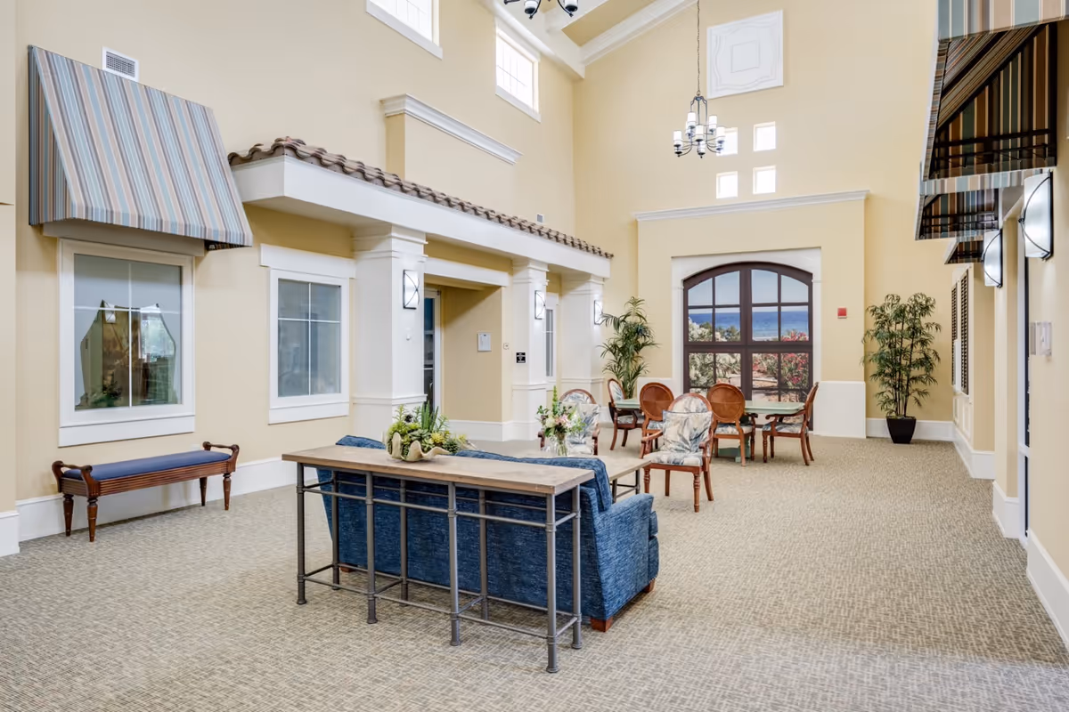 A spacious and well-lit common area with high ceilings, beige walls, and carpeted floor. The room features a blue sofa with a wooden console table behind it, several wooden chairs with cushions, a round table, potted plants, and large windows with striped awnings. A chandelier hangs from the ceiling, and a large window at the far end shows a scenic view of trees and water.