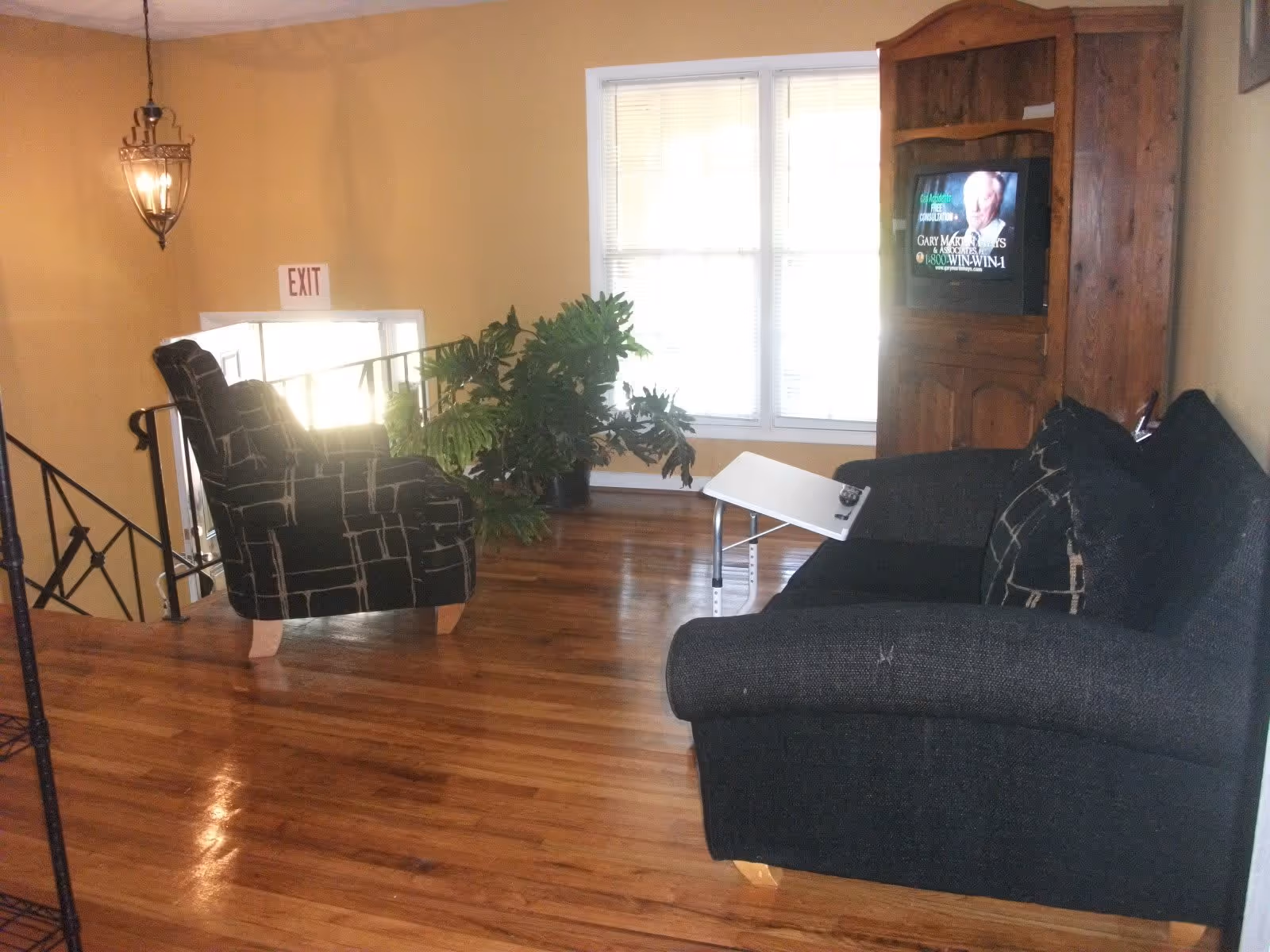 A cozy living room area with wooden flooring, a black patterned armchair, a black sofa with matching cushions, a small adjustable table with a remote control on it, a large green potted plant near a window with blinds, a wooden cabinet with a TV showing a commercial, and an exit sign above a door near a staircase.