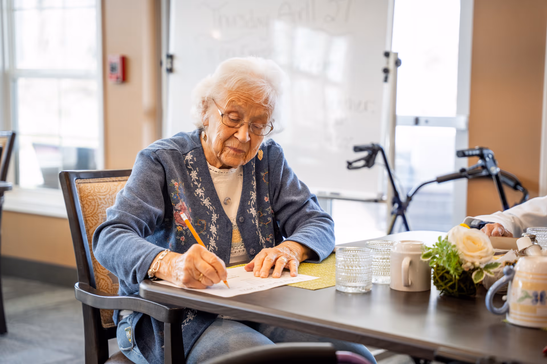 An elderly woman with white hair and glasses is sitting at a table in a well-lit room, writing on a piece of paper with a pencil. She is wearing a blue cardigan with floral embroidery and a white shirt underneath. On the table are several glasses, a white mug, and a small floral arrangement. In the background, there is a whiteboard and a walker near a window.