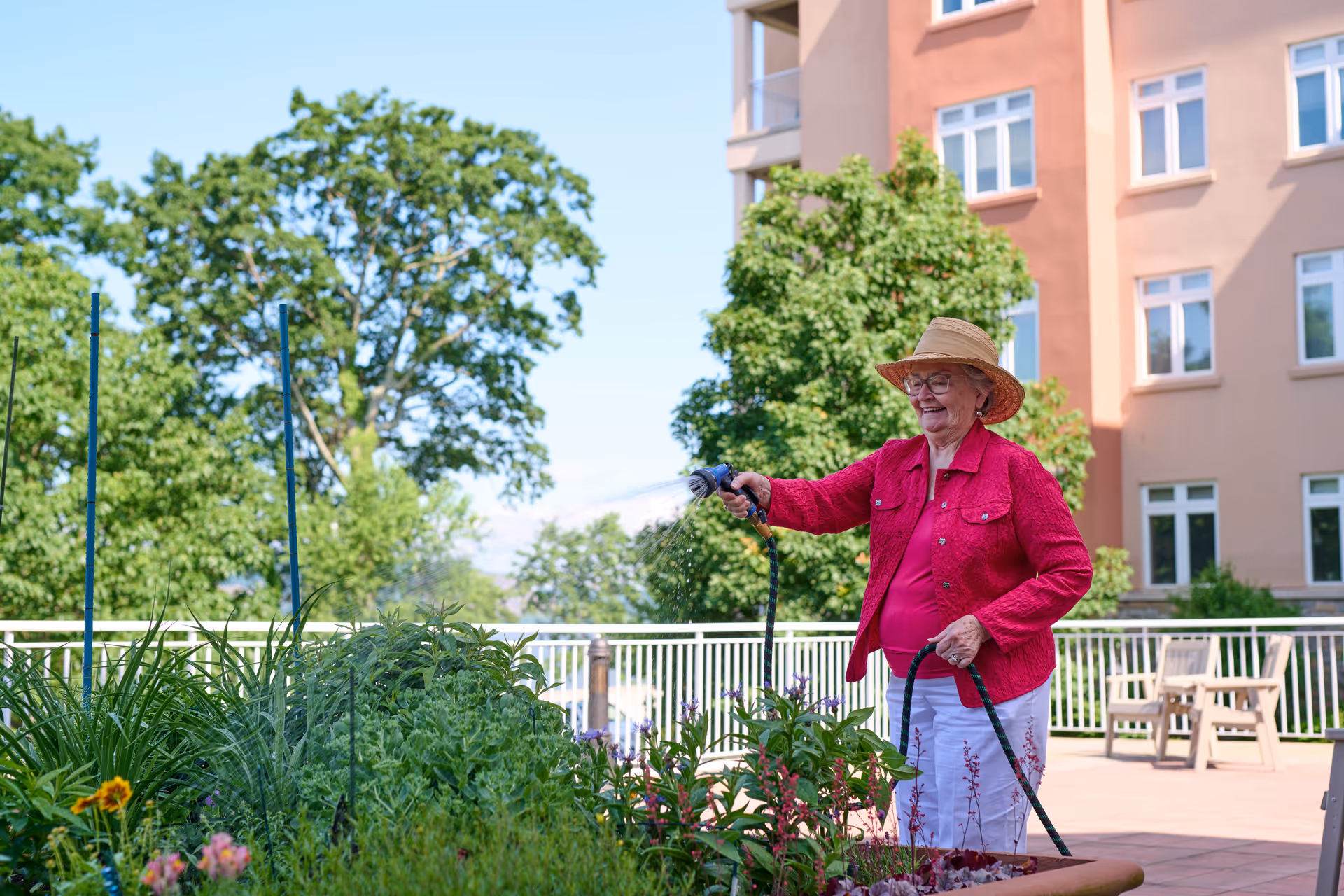 An elderly woman wearing a straw hat and a red jacket waters plants in a garden area outside a multi-story building on a sunny day.
