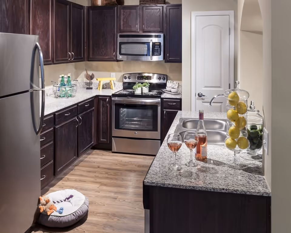 Modern kitchen with dark wood cabinets, stainless steel refrigerator, oven, and microwave. Granite countertops hold a bottle of rosé wine with two filled wine glasses, a glass container with lemons, and another container with limes. Wooden flooring and a small pet bed with toys are visible on the floor near the refrigerator.