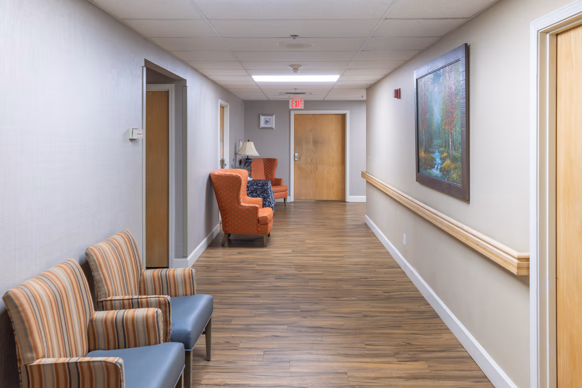 A clean, well-lit hallway in a senior living facility with wooden flooring and beige walls. There are several chairs along the walls, including two striped armchairs on the left and two orange armchairs further down the hall. A framed painting of a forest scene hangs on the right wall above a wooden handrail. A wooden door is visible at the end of the hallway beneath an illuminated exit sign.
