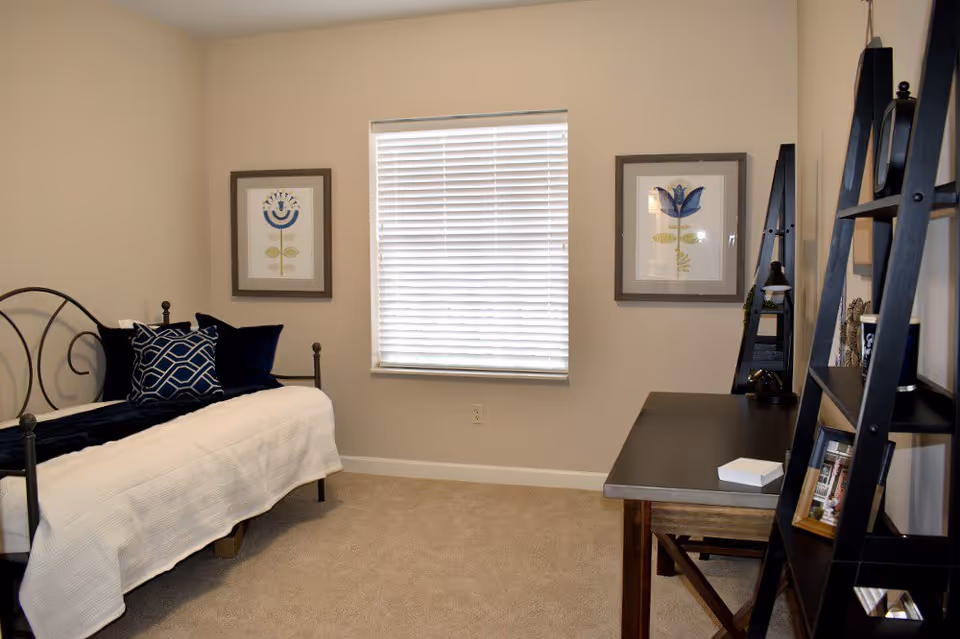 A neat bedroom with a daybed, decorative pillows, a window with blinds, and a desk with shelving.
