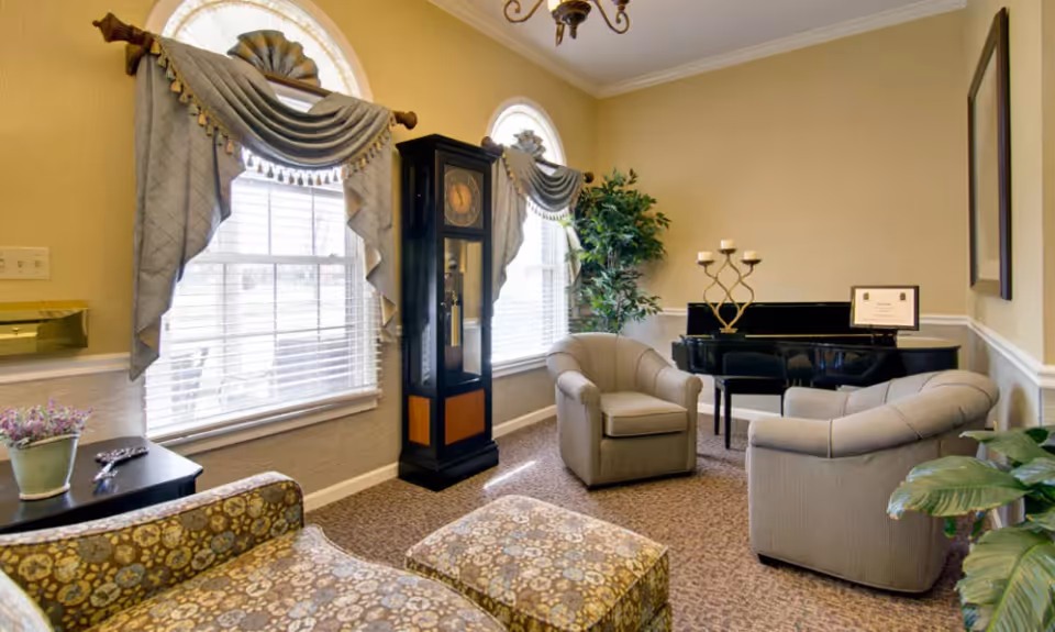 A cozy living room area with two large windows covered by decorative curtains, a tall grandfather clock between the windows, two upholstered armchairs, a patterned ottoman, a black grand piano with a candle holder and a framed certificate on top, and several potted plants. The walls are painted beige with white trim, and the floor is carpeted.