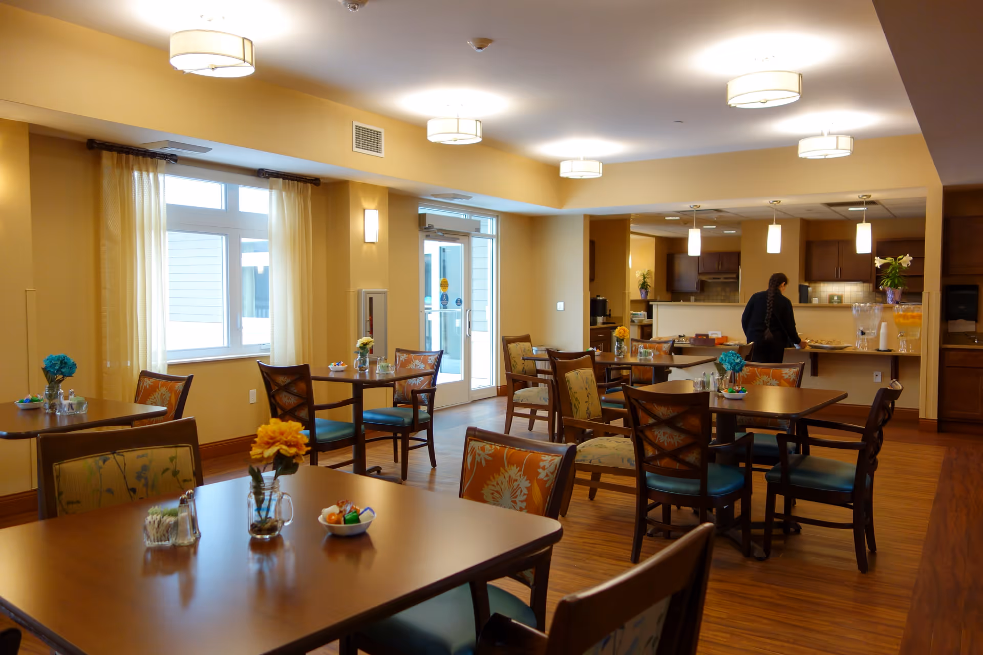 A cozy dining area in a senior living facility with several wooden tables and chairs, each table decorated with small flower vases and bowls of candy. The room has warm yellow walls, large windows with sheer curtains, and ceiling lights. In the background, a person is seen standing near a counter with beverages and snacks.