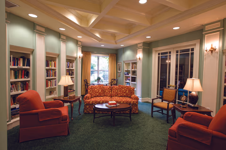 Cozy common room with a patterned sofa and armchairs around a coffee table, lamps, built-in bookshelves, and a window letting in daylight.