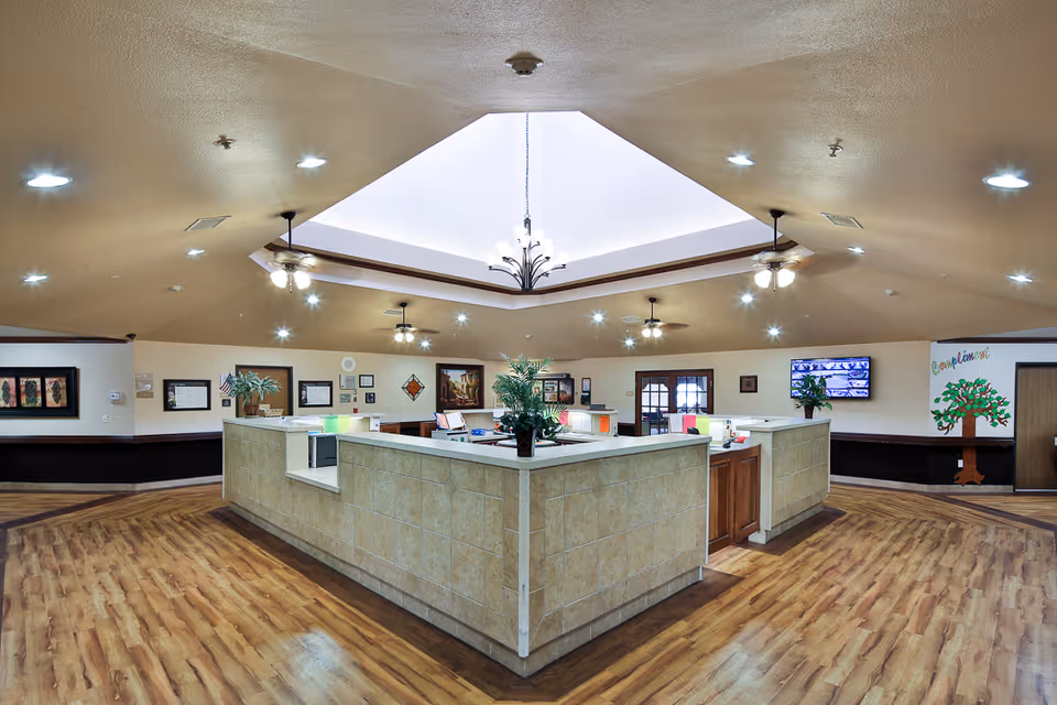 Interior view of a nursing and rehabilitation center reception area with a large, square-shaped front desk in the center. The ceiling has a recessed, octagonal skylight with a chandelier hanging down. The room is well-lit with multiple ceiling lights and ceiling fans. The floor is wooden, and the walls are decorated with framed pictures, plants, and a colorful welcome tree mural. A TV screen is mounted on the wall to the right.