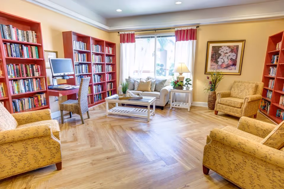A cozy living room area with light wooden flooring, beige walls, and large windows with red and white curtains. The room features multiple yellow patterned armchairs, a beige sofa with cushions, a white coffee table, and two white side tables with lamps and plants. There are red bookshelves filled with books along the walls, and a computer workstation with a chair is set up against one wall. A framed floral painting hangs on the wall.