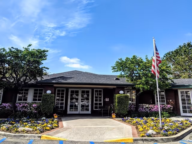 Entrance of a single-story brick building with a dark shingled roof, flanked by well-maintained flower beds with colorful flowers and green shrubs. An American flag on a flagpole stands to the right of the entrance under a clear blue sky.