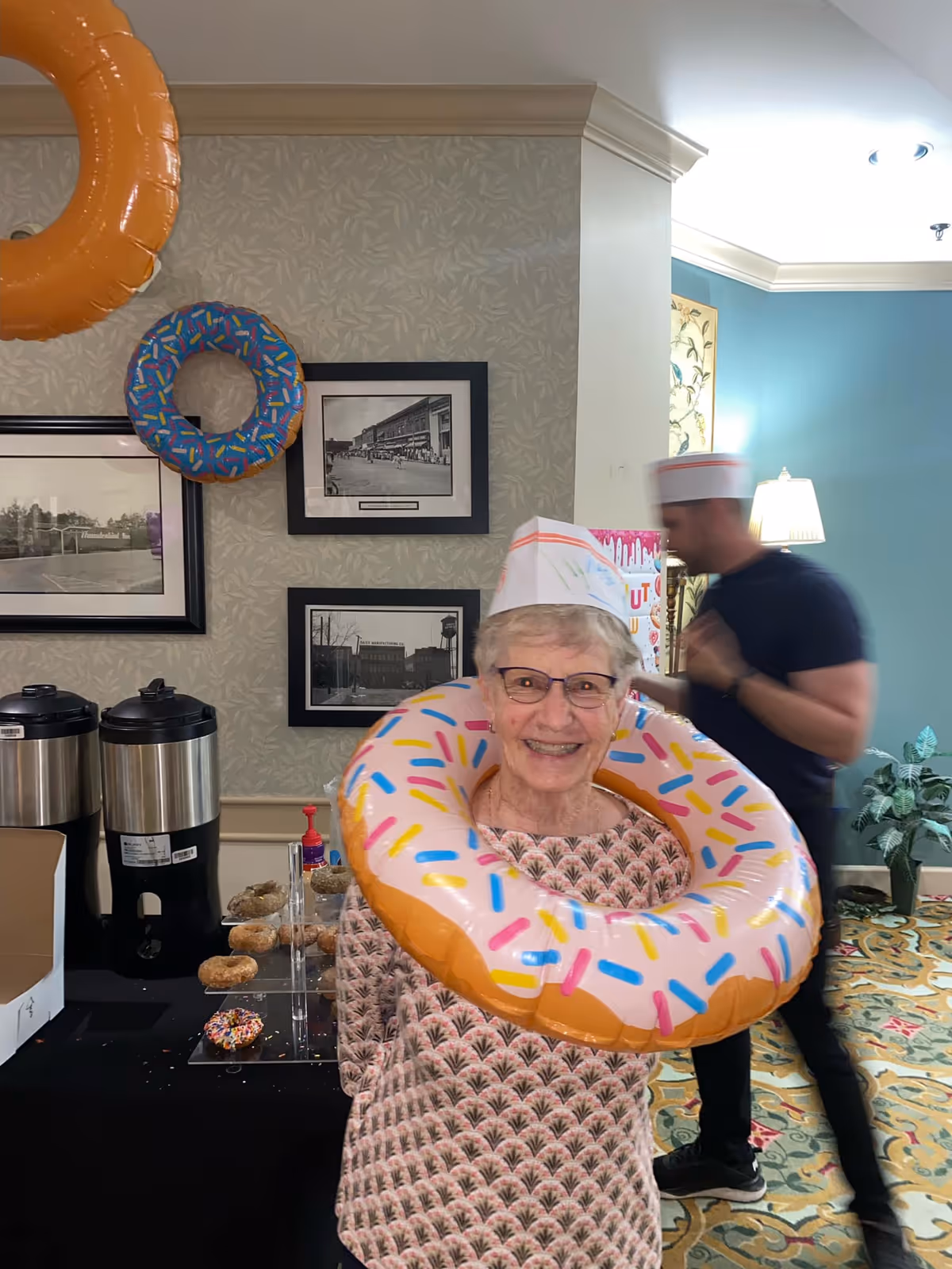 An elderly woman wearing glasses and a paper hat is smiling and posing with an inflatable donut around her neck in a decorated room. Behind her, there is a man also wearing a paper hat, a table with coffee dispensers, donuts, and a donut-themed decoration on the wall. The room has framed black and white photos on the wall and a patterned carpet.