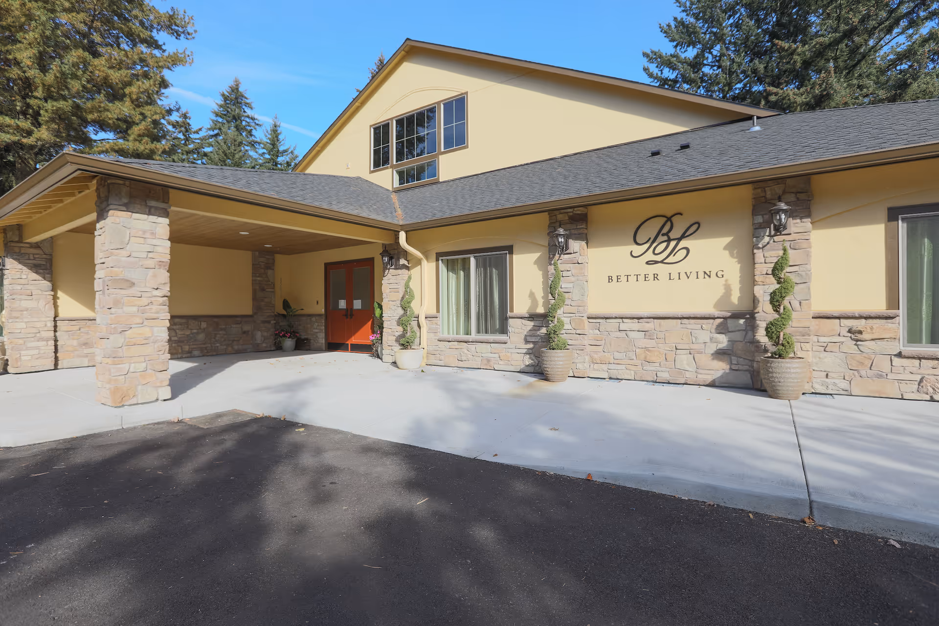 Exterior front view of the Better Living RCF facility showing a beige building with stone accents, a covered entrance supported by stone pillars, red double doors, potted plants, and the Better Living logo on the wall.