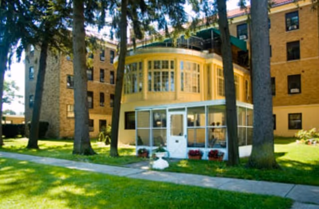 Brick multi-story senior living building with a rounded sunroom and screened porch, set behind a lawn and tall trees.