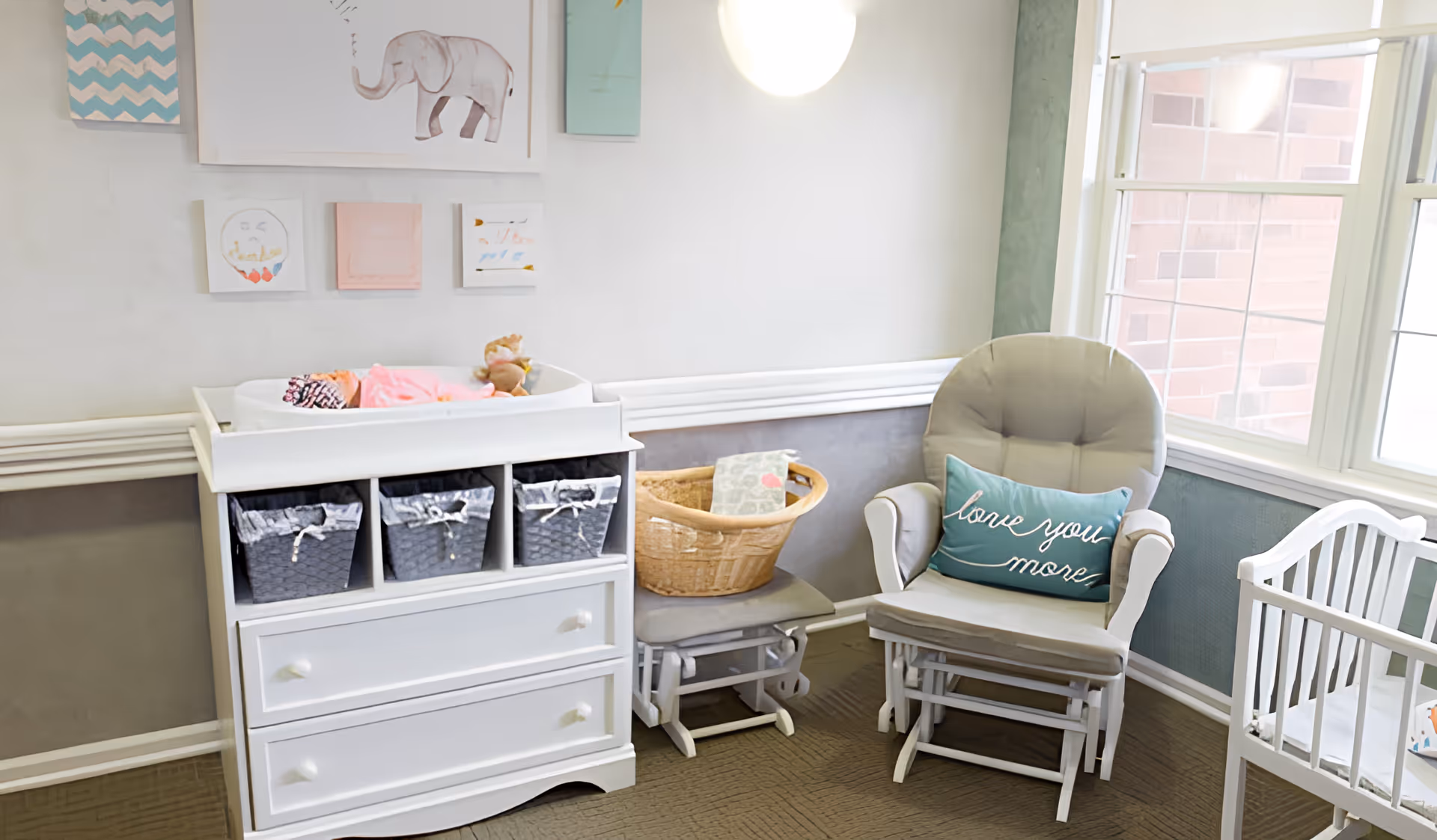 A bright nursery room featuring a white changing table with three woven baskets, a baby doll lying on top, a wicker basket on a small bench, a cushioned rocking chair with a blue pillow that says 'love you more', and a white crib near a window with natural light coming in. The walls are decorated with framed pictures including an elephant illustration.