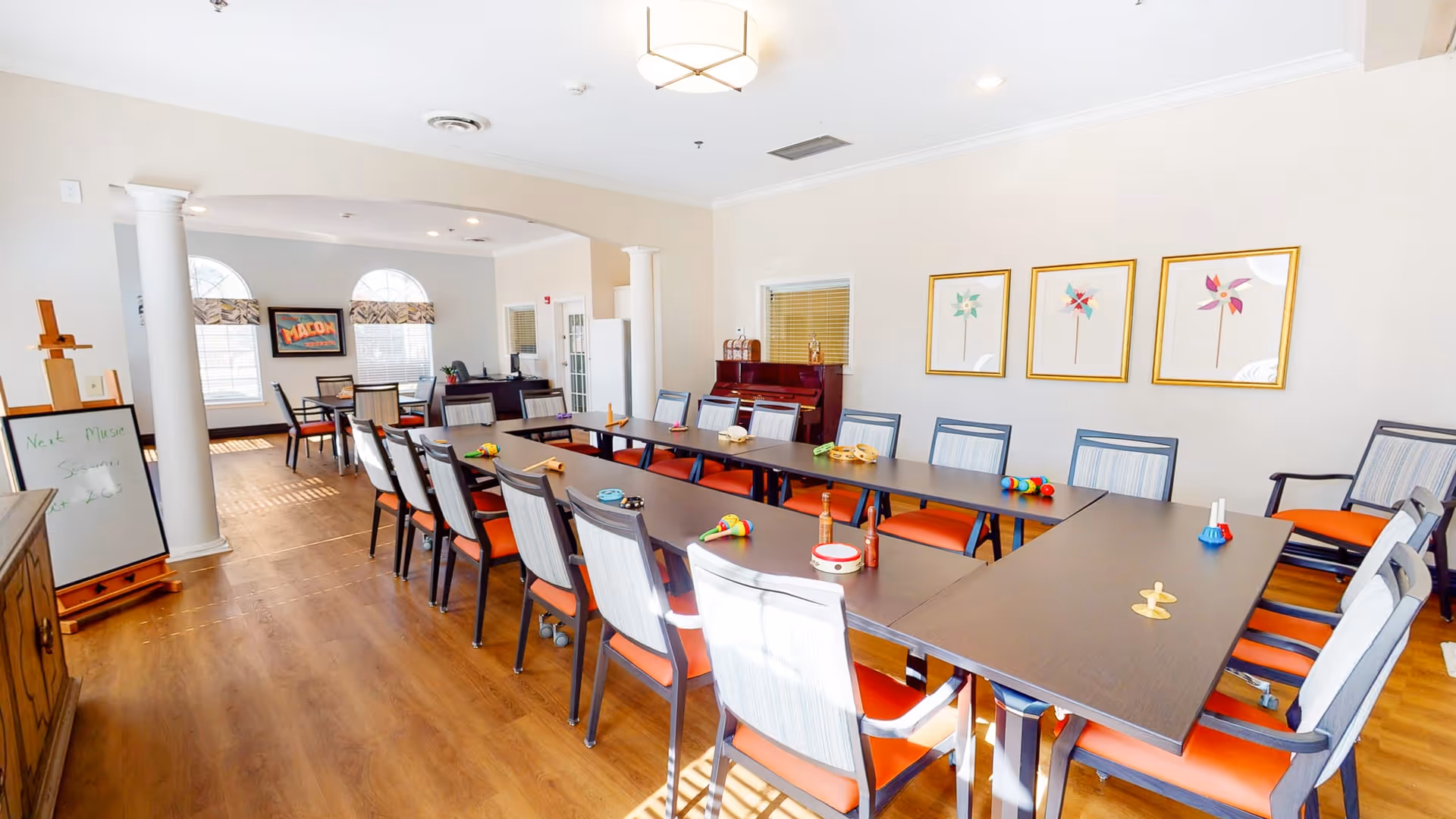 A bright and spacious activity room with a large U-shaped table surrounded by chairs with orange cushions. Various small musical instruments and toys are placed on the table. The room has wooden flooring, white walls, and three framed pictures of colorful pinwheels on one wall. There is a piano against the wall and a whiteboard on an easel near the entrance. Large windows with arched tops allow natural light to fill the room.