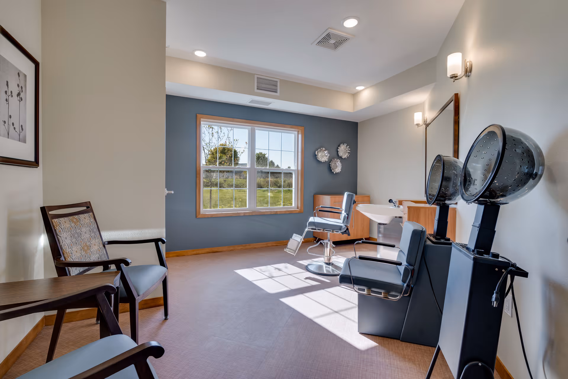 Interior of a senior living facility hair salon with two salon chairs, two hair dryers, a large window letting in natural light, a framed artwork on the wall, and two waiting chairs with a small table.