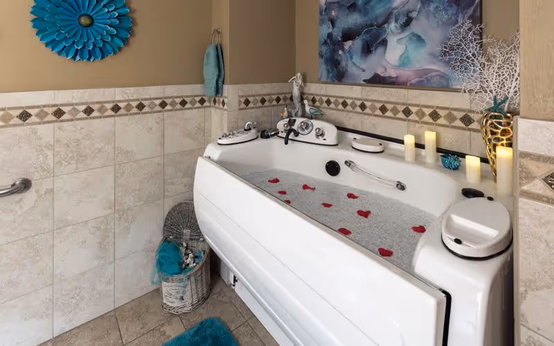 A luxurious bathtub filled with water and rose petals in a tiled bathroom. The tub is surrounded by decorative items including candles, a blue flower wall decoration, a basket with towels, and a piece of abstract art on the wall.