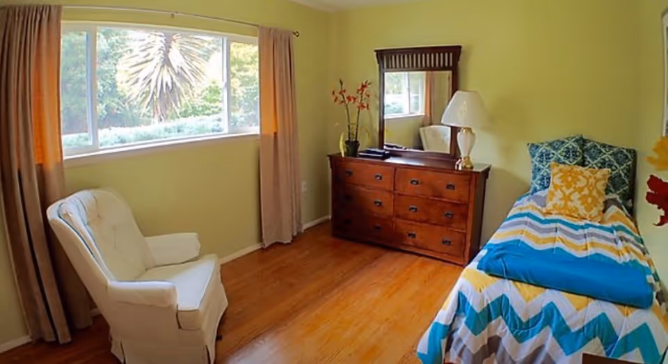 A bright bedroom with light green walls and wooden flooring. The room features a single bed with a colorful chevron-patterned bedspread in blue, yellow, and white, accompanied by two decorative pillows. Next to the bed is a wooden dresser with a mirror, a table lamp, and a small plant with orange flowers. A white upholstered armchair is placed near a large window with beige curtains, allowing natural light to fill the room.