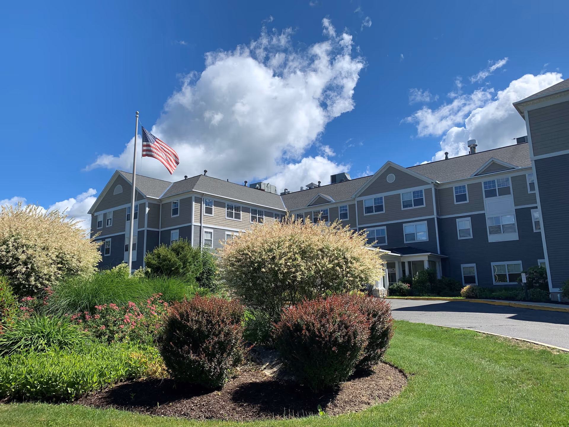 Exterior view of a multi-story senior living facility building with beige and gray siding under a blue sky with scattered clouds. In front of the building, there is a landscaped area with various bushes and plants, and an American flag on a flagpole.