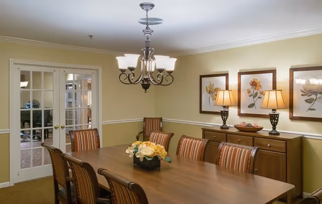 A dining room with a long wooden table surrounded by eight striped upholstered chairs. On the table is a floral centerpiece. The walls are painted light yellow with white trim, and three framed floral artworks hang above a wooden sideboard with two table lamps and a decorative bowl. A chandelier with five lights hangs above the table. French doors with glass panes are visible in the background.