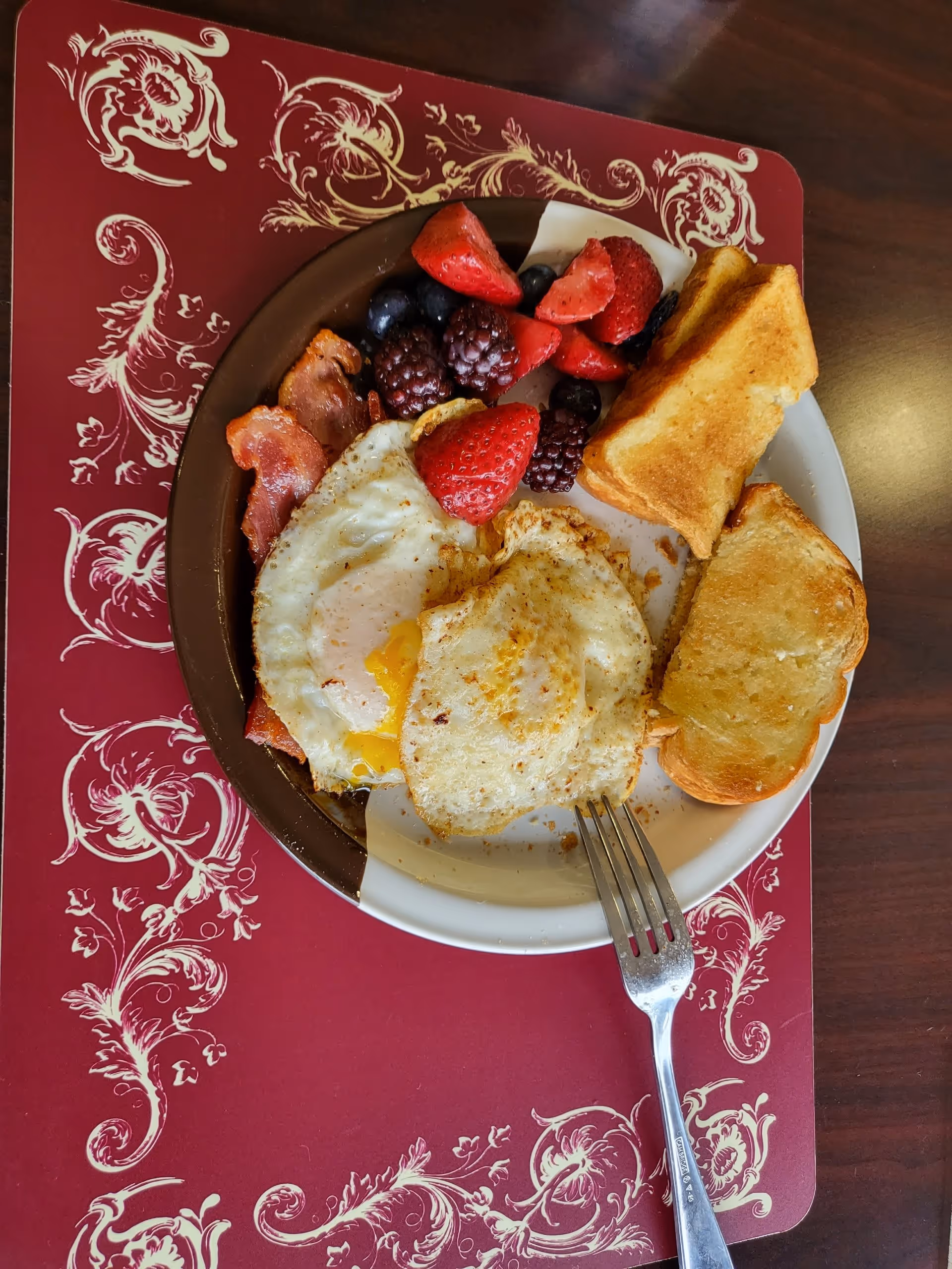 A plate with two fried eggs, two slices of toasted bread, several pieces of bacon, and a mix of fresh berries including strawberries, blackberries, and blueberries on a decorative red placemat with cream-colored floral patterns. A fork rests on the plate.