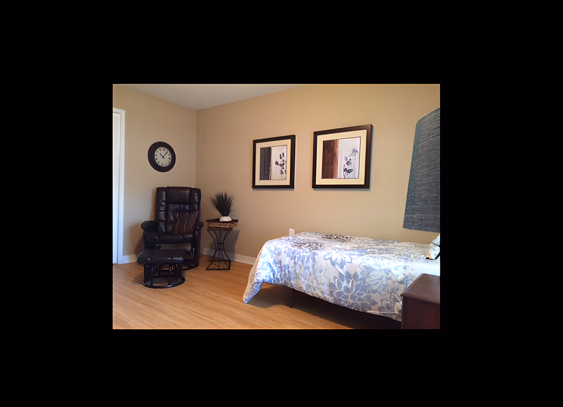 A cozy bedroom with a single bed covered in a floral-patterned blanket, a dark leather recliner chair with a matching footrest, a small side table with decorative items, two framed artworks on the beige wall, a wall clock, and a lamp partially visible on the right side.