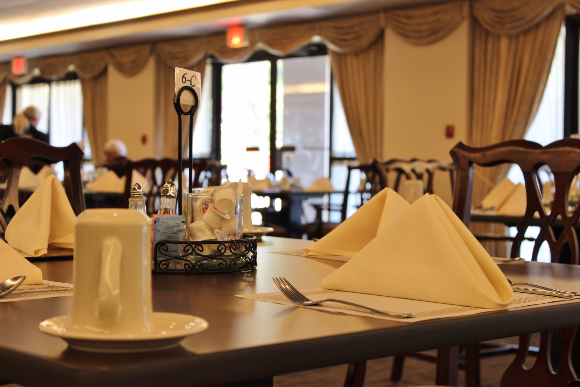 A dining table set with folded cream-colored napkins, silverware, a white cup on a saucer, and a condiment holder with salt, pepper, sugar packets, and creamers. The background shows more tables and chairs with similar settings and large windows with curtains.