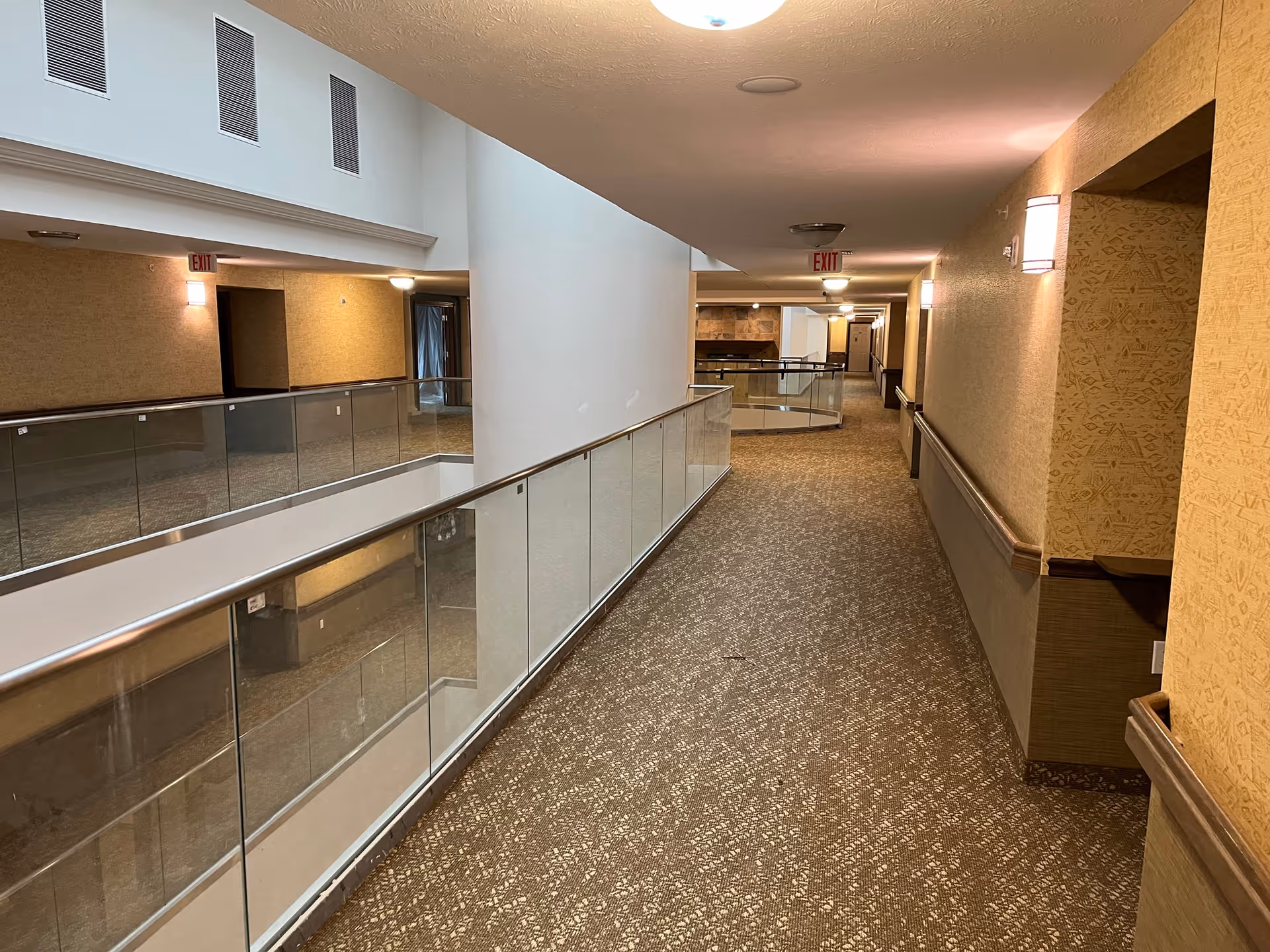 Carpeted indoor hallway with glass railing overlooking an atrium, wall sconces, and exit signs in a retirement community.