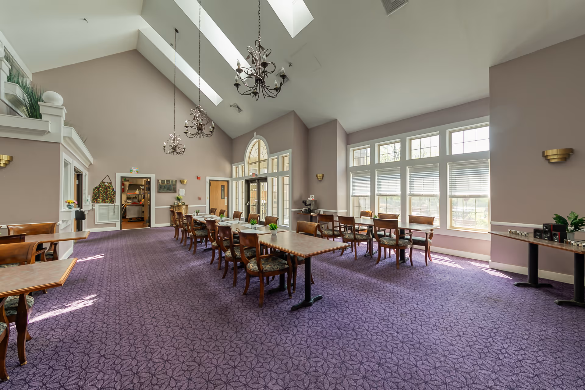 Spacious dining room with long wooden tables and chairs, large windows and skylights, chandeliers, and purple patterned carpet.