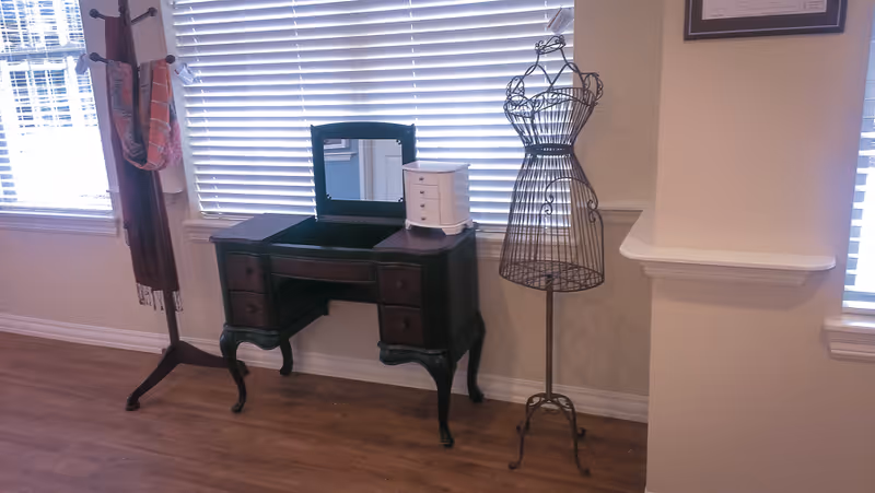 A wooden vanity desk, coat rack and wire dress form stand against a wall with closed blinds over hardwood floors.