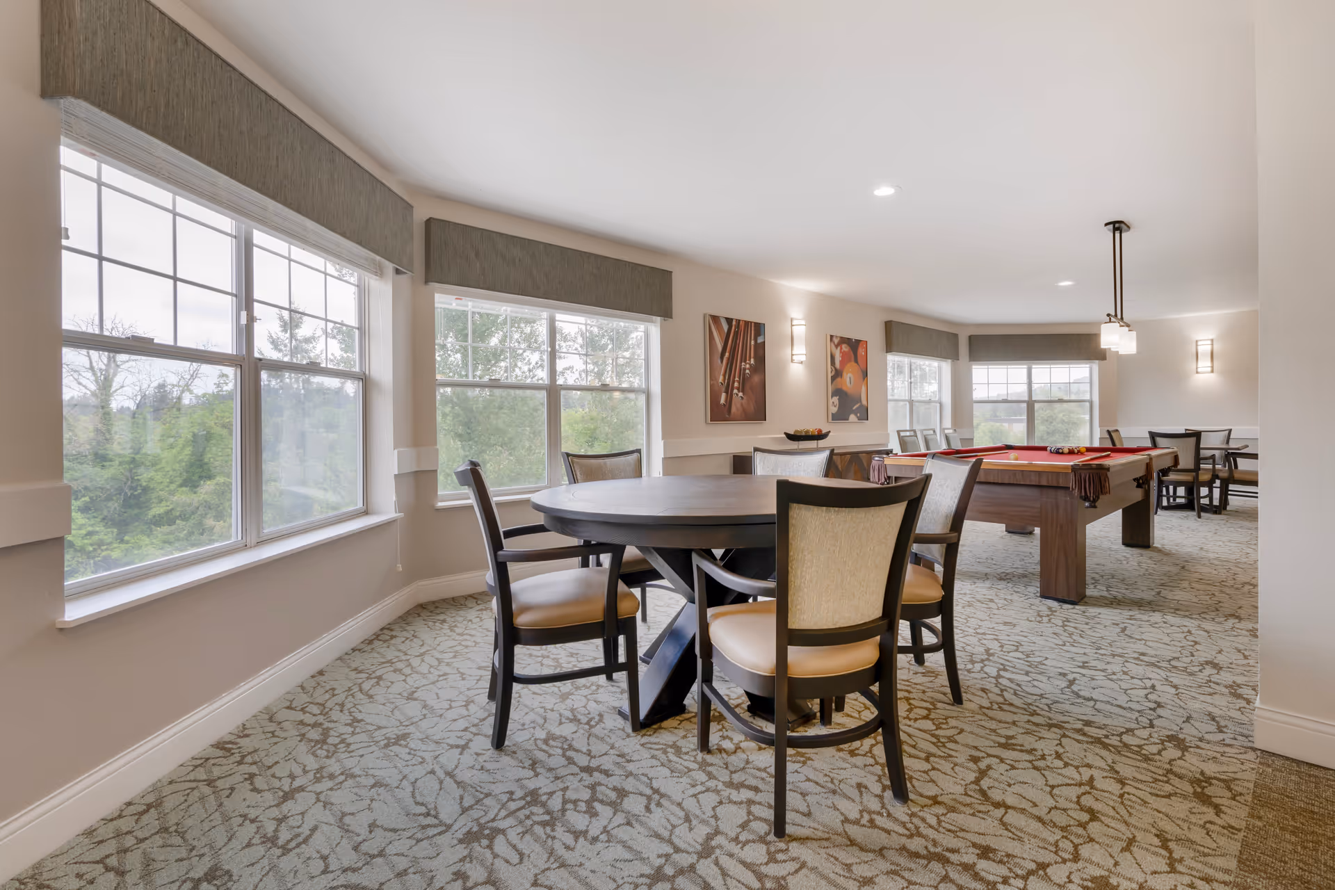 A bright and spacious common area with large windows showing greenery outside. The room features a round table with four chairs in the foreground and a pool table with a red felt surface in the background. There are additional tables and chairs near the windows, and framed artwork on the walls. The carpet has a subtle leaf pattern, and the lighting is soft with wall sconces and a hanging light fixture above the pool table.