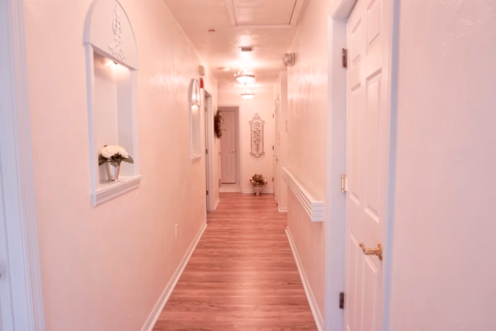 Long softly lit interior hallway with wood flooring, doors and decorative wall niches in an assisted living facility.