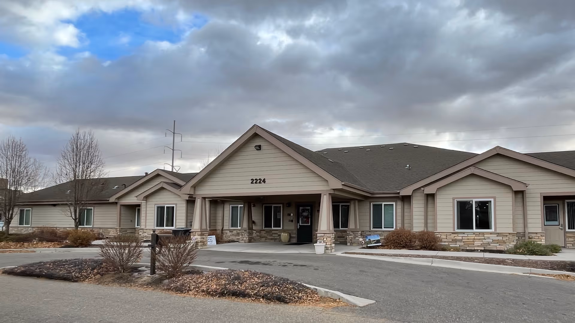 Exterior view of a single-story senior living facility building with beige siding and stone accents, a covered entrance with the number 2224 above it, and a cloudy sky overhead.