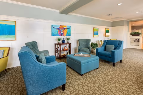 A cozy seating area in a senior living facility with four blue upholstered armchairs arranged around a square blue ottoman. The room has patterned carpet flooring, white paneled walls with a light green upper section, and colorful artwork including a painting of a bull and landscape paintings. There is a small wooden shelf with decorative items and plants, and a lamp on a side table in the background.