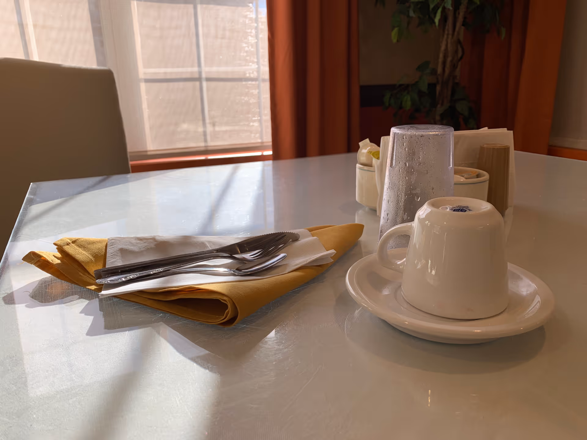 A dining table set with a folded yellow napkin, a white napkin, a fork and spoon placed on top. There is an upside-down white coffee cup on a saucer, a glass of water with condensation, and a container holding sugar packets and salt and pepper shakers. Sunlight is coming through a window with curtains in the background.