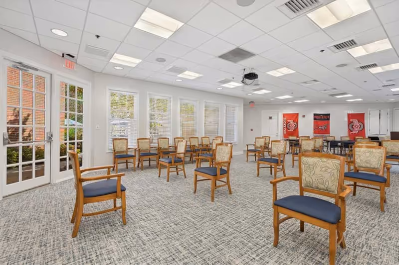 A spacious, well-lit room with multiple wooden chairs arranged in rows, each chair having blue cushioned seats and patterned backs. The room has large windows with white blinds, a glass door leading outside, and a carpeted floor. The ceiling has recessed lighting and a projector mounted in the center. Red banners are visible on the far wall.