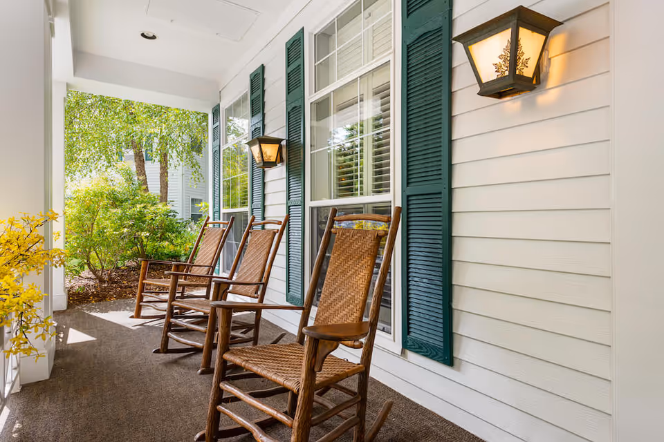 Covered exterior porch with a row of wooden rocking chairs in front of windows with green shutters and wall lanterns overlooking landscaped greenery.