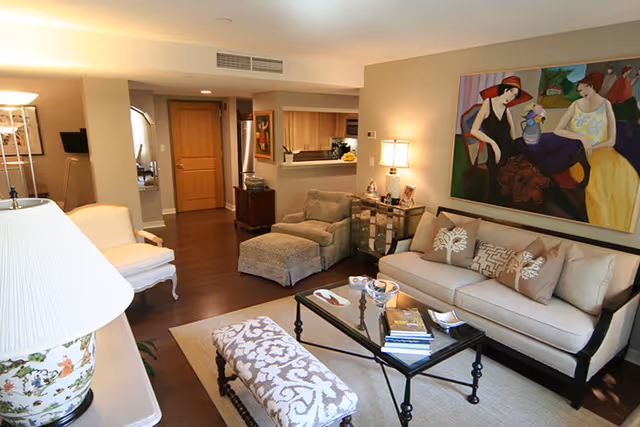 A cozy living room with a beige sofa adorned with patterned pillows, a glass coffee table with books and decorative items, an upholstered bench with a floral pattern, and a comfortable armchair with an ottoman. The room features warm lighting from a table lamp and a floor lamp, wooden flooring, and a colorful painting of two women on the wall. The background shows a kitchen area and a wooden door.