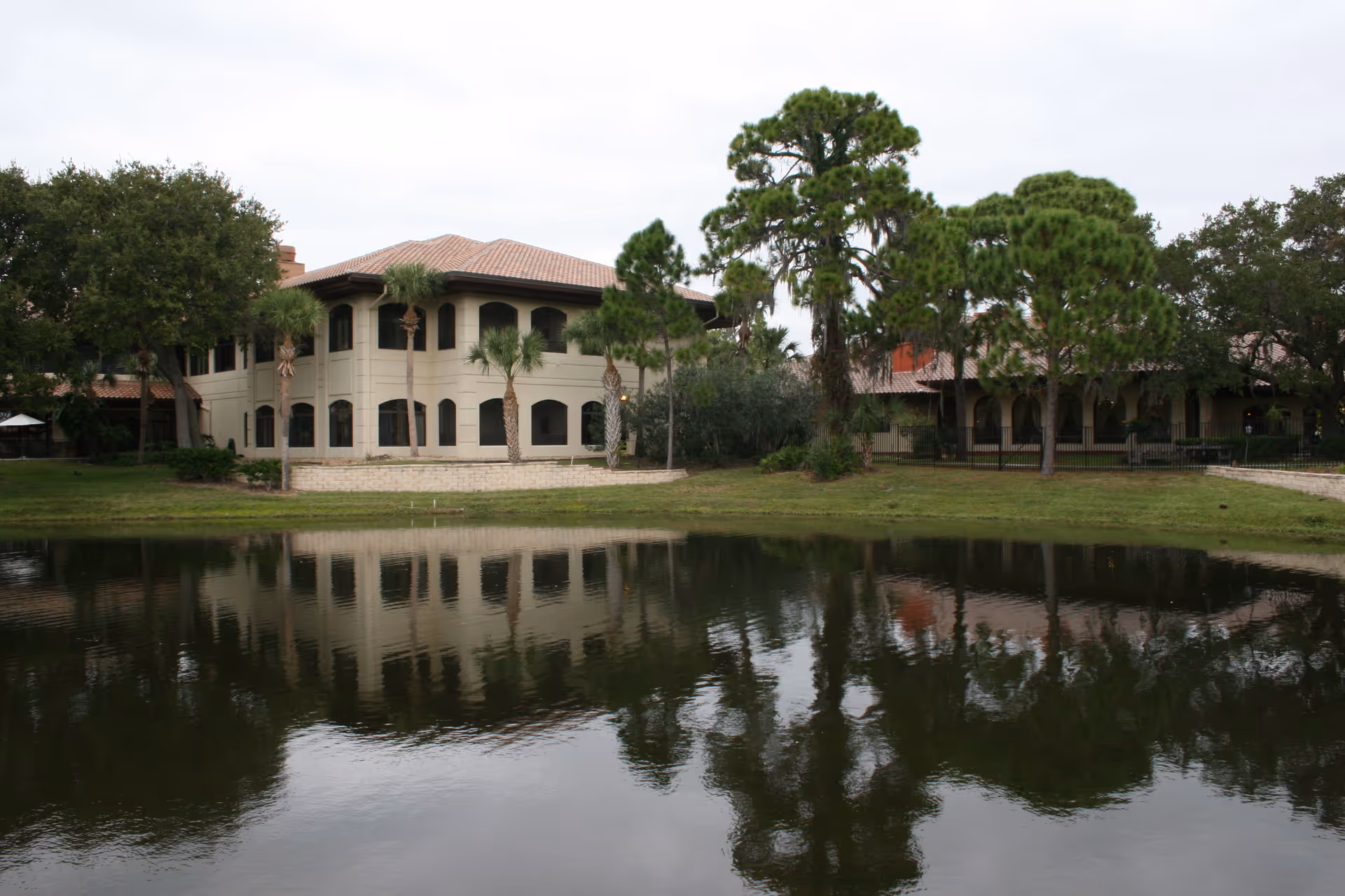 Two-story Mediterranean-style building with arched windows and palm trees reflected in a pond.