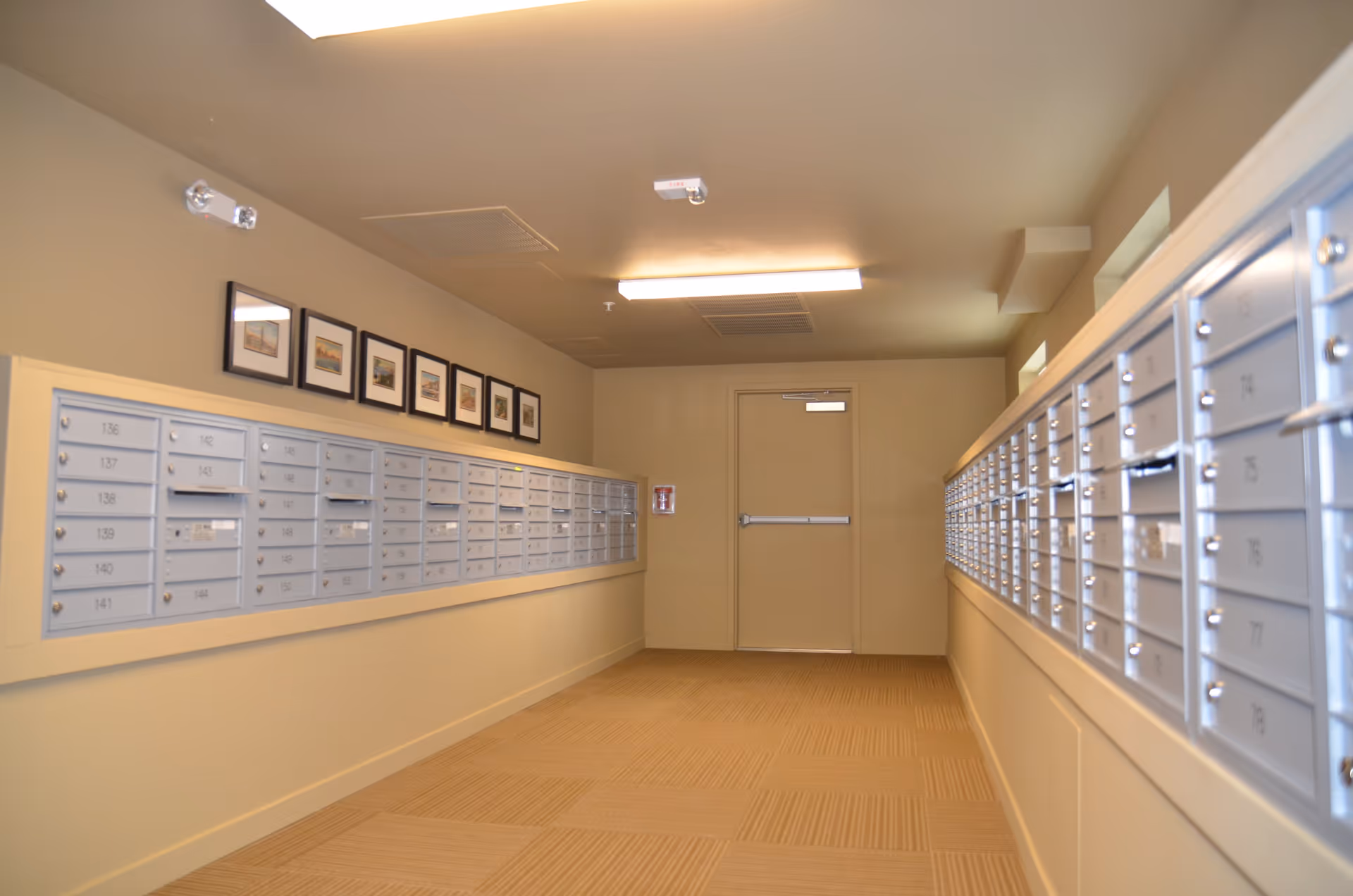 A hallway with rows of metal mailboxes mounted on both walls, beige carpeted floor, beige walls, a closed door at the end, and framed pictures hanging above the mailboxes on the left wall.