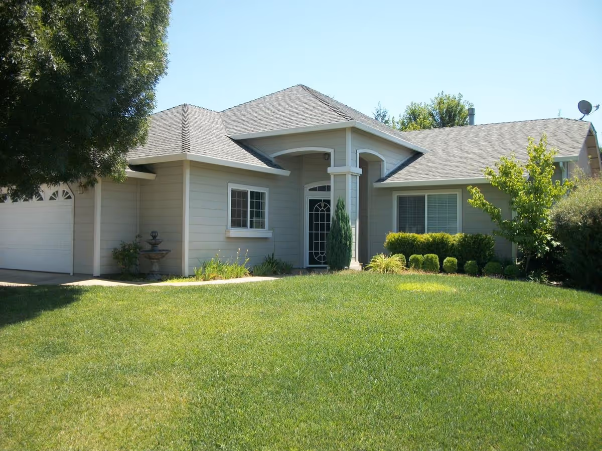 Single-story house with a gray exterior, a front door with decorative glass, a garage on the left, and a well-maintained green lawn with shrubs and trees under a clear blue sky.