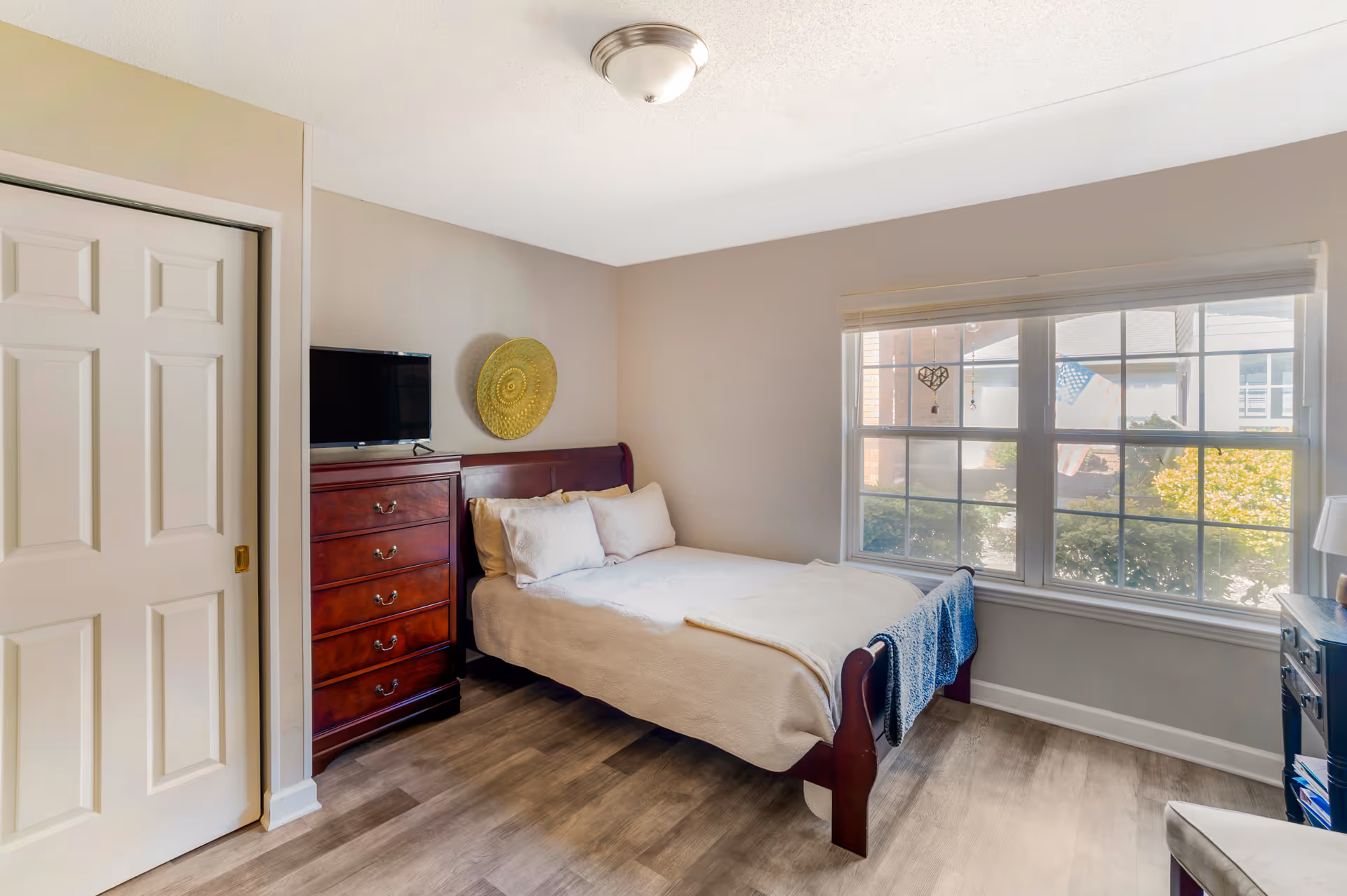 A bright bedroom with a wooden bed frame and white bedding, a wooden dresser with a flat-screen TV on top, a large window with a view of greenery and an American flag outside, and light-colored walls and flooring.