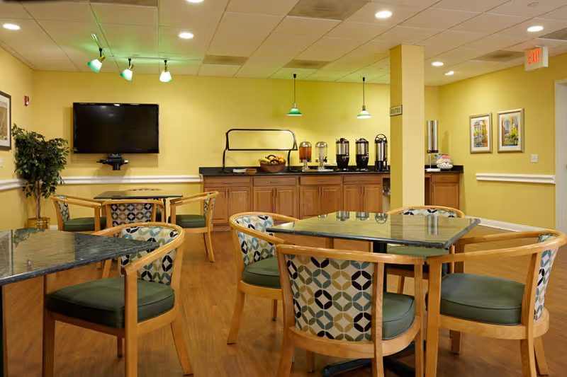 A dining area in a senior living facility with several tables and chairs. The chairs have wooden frames and patterned upholstery with green cushions. There is a counter against the wall with beverage dispensers and a basket of fruit. A flat-screen TV is mounted on the wall, and the room is lit by ceiling lights and green pendant lamps. The walls are painted yellow, and there are framed pictures and a potted plant in the corner.