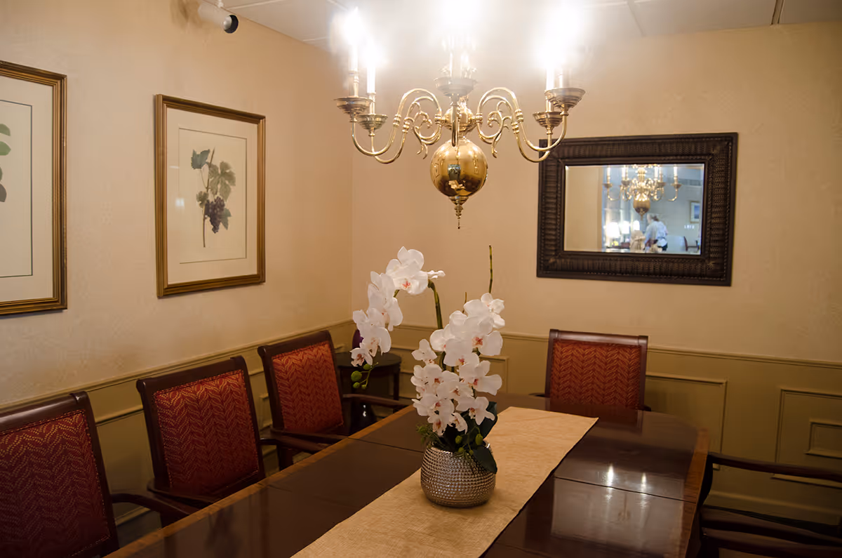 A dining room with a polished wooden table, six red cushioned chairs, a beige table runner, and a vase with white orchids in the center. The room has beige walls with framed botanical prints and a large mirror reflecting a chandelier and part of the room.