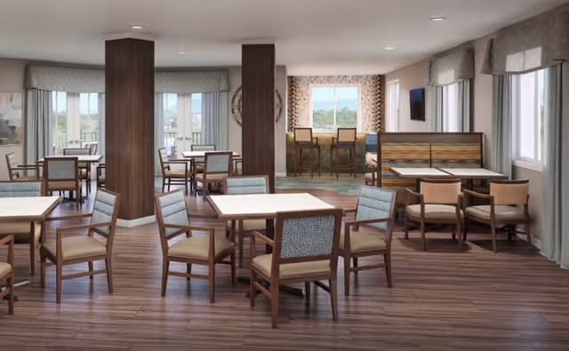 A spacious dining room with multiple square tables and wooden chairs with cushioned seats and backs. The room has large windows with curtains allowing natural light to enter. There are two wooden columns in the center, and a seating area with a colorful cushioned bench along one wall. The floor is wood, and the walls are light-colored with some patterned wallpaper near the windows.