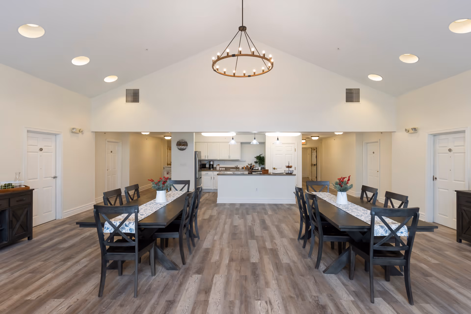 A spacious dining area with two long dark wooden tables, each surrounded by six matching chairs. Each table has a white vase with red flowers and a patterned runner. The room has light-colored walls, wood flooring, and a high vaulted ceiling with a circular chandelier. In the background, there is a kitchen area with white cabinets, a refrigerator, and pendant lights. Several white doors line the walls on either side of the room.