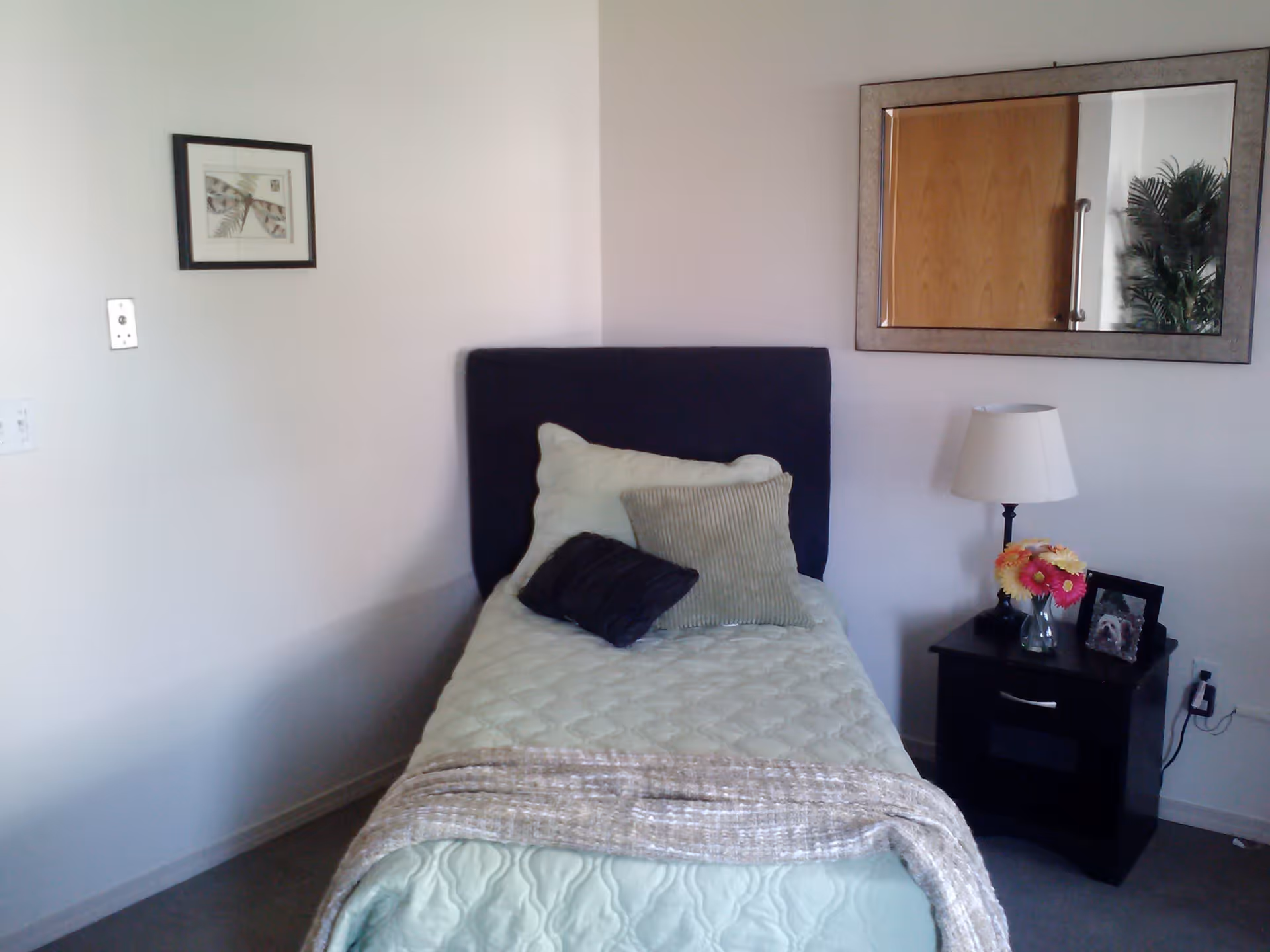 A small bedroom with a single bed covered in a light green quilt and three pillows. Next to the bed is a black nightstand with a lamp, a vase of flowers, and a framed photo. A large rectangular mirror hangs on the wall above the nightstand, reflecting a door and a plant. A framed picture is hung on the adjacent wall.