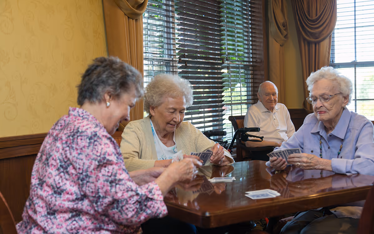 Four elderly individuals sitting around a wooden table playing cards in a room with large windows and yellow patterned walls. Three women are actively holding cards, while an elderly man sits in the background watching. The room has brown curtains and wooden paneling.